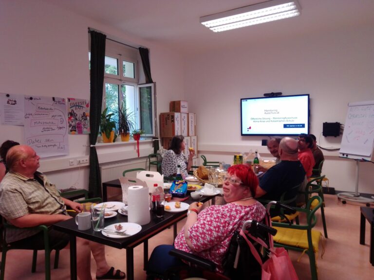 Personen sitzen gemeinsam ein einem Tisch auf dem Essen und trinken steht. Im Hintergrund ist ein Bildschirm auf dem der live Stream des Monitoring Ausschuss läuft.